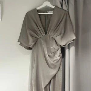 Ljusgrå långklänning från ASOS Edition - Elegant ljusgrå långklänning med djup v-ringning och vida korta ärmar. Klänningen har draperad detalj framtill och är tillverkad i ett glansigt, satinliknande material som ger en lyxig känsla. Perfekt för balen eller annat🥰Den är typ champange silver färgad.