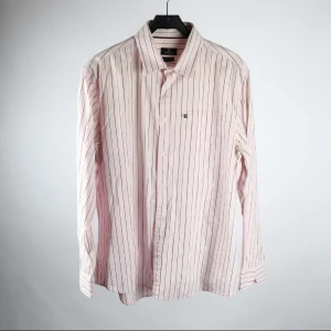 Randig skjorta från U.S. Polo Assn. - Säljer en klassisk ljusrosa skjorta från U.S. Polo Assn. med tunna blå och vita ränder. Skjortan har lång ärm, knappar framtill och en bröstficka. Tillverkad i 100% bomull för en skön känsla.