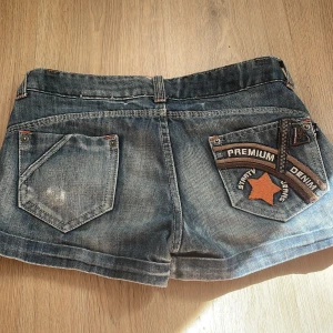 Blå jeansshorts med stjärndetalj - Säljer ett par blå jeansshorts med cool stjärndetalj och texten 'Premium Denim' på ena bakfickan. Klassisk femficksmodell med knapp och dragkedja framtill. Perfekta för sommaren!