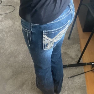 Jeans - Säljer ett par blå, lågmidjade jeans med snygga broderade detaljer och paljetter på bakfickorna från Gina Tricot. Jeansen har vita kontrastsömmar och klassisk femficksdesign. Passformen är bootcut och materialet är denim.