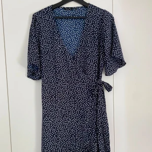 Marinblå omlottklänning med vita prickar - Säljer en marinblå omlottklänning med vita prickar och volang nertill. Klänningen har korta ärmar och knyt i midjan för en snygg passform. 