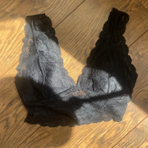Svart spets bralette - Säljer en svart bralette med v-ringning och breda spetskantade axelband. Toppen är helt i spets och har en mjuk, feminin look. Perfekt att styla under en skjorta eller kavaj.