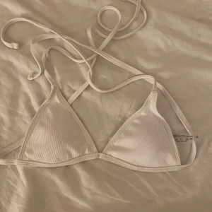 Vit ribbad trekantsbikini med knyt - Säljer en vit trekantsbikini i ribbat material. Bikinin har tunna band som knyts i nacken och ryggen för justerbar passform. Enkel och stilren design, perfekt för sommaren. Den är aldrig använd och säljer den för att den är för liten för mig!