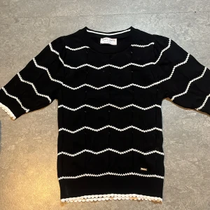 Svart stickad topp från Holly & Whyte - Svart stickad topp från Holly & Whyte med vita sicksack-ränder och dekorativa kanter. Tröjan har rund halsringning och halv-långa ärmar. Perfekt för dig som gillar en klassisk men lekfull stil.