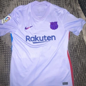 FC Barcelona bortatröja Gavi 30 - Säljer en lila FC Barcelona bortatröja från Nike med Gavi och nummer 30 på ryggen. Tröjan har korta ärmar, Rakuten-tryck på bröstet och La Liga-märke på ärmen. Materialet är Dri-FIT för bra andningsförmåga. Perfekt för dig som älskar Barca! Hör av er vid funderingar😁
