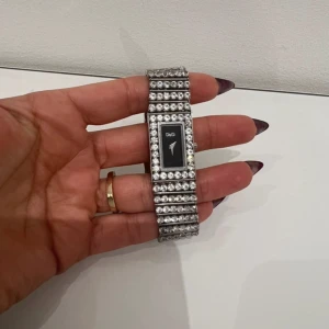 Glittrig klocka från D&G - Säljer en riktigt snygg och glittrig klocka från D&G med rektangulär svart urtavla och silverfärgat armband täckt av gnistrande stenar. Perfekt accessoar för dig som vill sticka ut och addera lite extra bling till din stil.