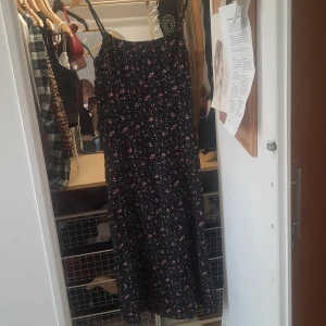 Svart blommig långklänning med rosett - Svart långklänning med smala axelband och små rosa blommor över hela tyget. Klänningen har en söt knytrosett i midjan och ett luftigt fall. Perfekt för varma dagar eller sommarens tillfällen.