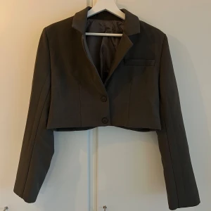 croppad kavaj - Säljer en stilren brun/grön croppad kavaj med två knappar framtill och krage. Aldrig använd och köptes från en butik i Polen förra året. Har även i svart som jag skulle kunna tänka mig att sälja om det är av intresse ☺️
