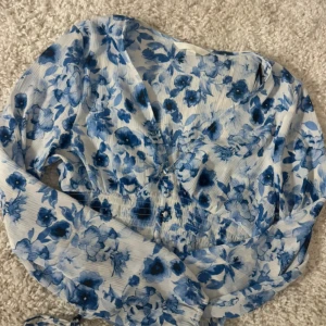 Blommig croppad blus i blått och vitt - Säljer en croppad blus med blått blommönster på vit botten. Blusen har långärmade puffärmar, v-ringning och smockad midja. Perfekt för vår och sommar!