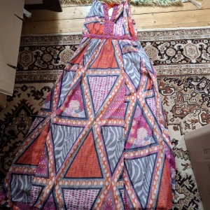 Färgglad långklänning med geometriskt mönster - Säljer en långklänning med färgstarkt geometriskt mönster i orange, lila, rosa och grått. Klänningen har smala axelband och v-ringning med dekorativa detaljer upptill. Perfekt för dig som gillar unika och iögonfallande plagg.