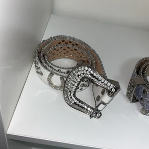 Glittrigt bälte med strass och nitar - Säljer ett snyggt bälte i silverfärg med massor av glittrande strass och små nitar längs hela bältet. Spännet är också täckt av strass för extra bling. Perfekt accessoar för att lyfta vilken outfit som helst.