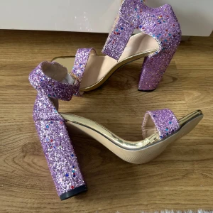 Lila glittriga klackskor med bred klack - Säljer ett par lila sandaler med bred klack och öppen tå. Skorna är täckta av glitter i olika färger och har en justerbar rem med spänne runt ankeln. Perfekta för att sticka ut och ge extra glans till outfiten. Oanvända storlek 36