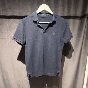   Ralph Lauren Piké - Klassisk blå pikétröja från Polo Ralph Lauren med korta ärmar och krage. Tröjan har två knappar vid halsen och den ikoniska broderade loggan på bröstet. Den har en fläck till vänster som man kan se på bilden, men går troligtvis bort i tvätt. Skriv vid minsta fundering! 