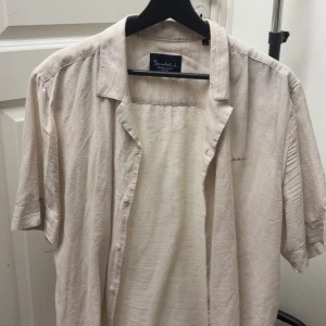 Beige linneskjorta från Bondelid - Säljer en stilren beige kortärmad skjorta från Bondelid. Skjortan är tillverkad i linne och har klassisk krage samt knäppning framtill. Perfekt för varma dagar och enkel att matcha med jeans eller shorts.