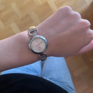 Silverfärgad analog klocka från Foga - Stilren analog klocka från Foga med rund vit urtavla och tydliga siffror. Klockan har ett silverfärgat metallarmband med elastisk länk, vilket gör den enkel att ta på och av. Perfekt accessoar för dig som gillar klassisk design.