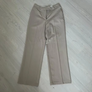 Beige kostymbyxor från NA-KD - Snygga beige kostymbyxor från NA-KD med raka och vida ben. Byxorna har pressveck framtill, hällor för bälte och stängs med knapp och dragkedja. Aldrig andvänts.