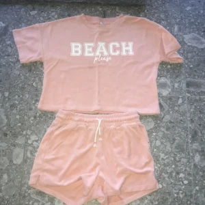 Rosa shorts från Lindex - Mjuka rosa shorts från Lindex med dragsko i midjan. Perfekta för sommaren och strandhäng. Matchar toppen med texten 'BEACH please'. Tillverkade i frottéliknande material.
