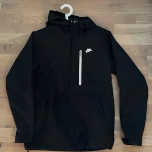 Nike vindjacka - Snygg svart vindjacka från Nike med huva och vit dragkedja på bröstfickan. Jackan har en stilren design med Nike-logga på bröstet och praktiska fickor. Perfekt för blåsiga dagar.