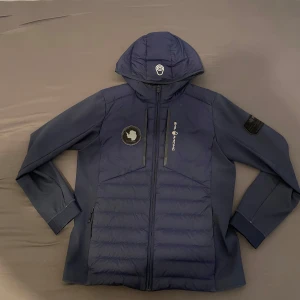 Blå Sail Racing JR Antarctica Hybrid Hood jacka - Säljer en blå Sail Racing JR Antarctica Hybrid Hood jacka med huva och dragkedja. Jackan har logga på bröstet och är tillverkad i en mix av nylon, polyester och dun. Snygg och sportig design med detaljerade märken på ärmen och bröstet.
