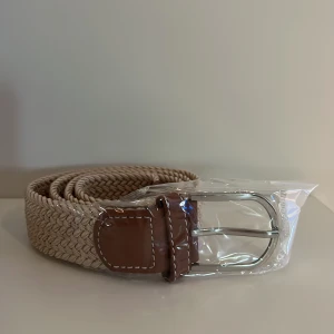 Beige flätat bälte  - Snyggt beige flätat bälte med detaljer i brunt läder och ett klassiskt silverfärgat spänne. Perfekt accessoar för att lyfta din outfit med en stilren touch. Passar både jeans och chinos.