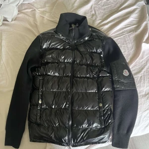 Svart pufferjacka från Moncler - Säljer en svart pufferjacka från Moncler med glansig framsida och stickade ärmar. Jackan har dragkedja framtill, hög krage och Moncler-logga på ärmen. Perfekt för kyliga dagar och snygg till många outfits.