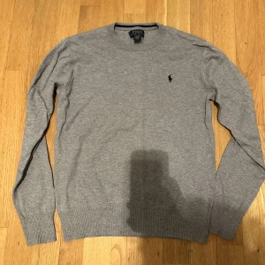 Grå tröja från Polo Ralph Lauren - Säljer en klassisk grå tröja från Polo Ralph Lauren med rund halsringning och broderad logga på bröstet. Tröjan har långa ärmar och ribbade muddar vid ärmslut och nederkant. Perfekt för en stilren look.