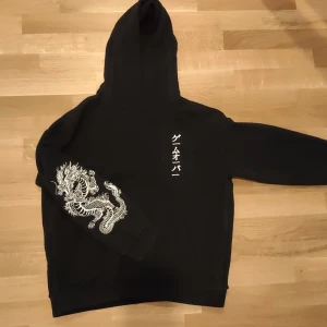 Svart hoodie med draktryck och japansk text - Säljer en svart hoodie med vit drake tryckt på ena ärmen och japansk text på bröstet. Hoodien har en klassisk huva och normal passform