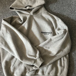 Grå hoodie från Essentials Fear of God - Säljer en grå hoodie från Essentials Fear of God med svart logga på bröstet och stor logga på ryggen. Klassisk känguruficka och huva. Perfekt för en avslappnad stil.