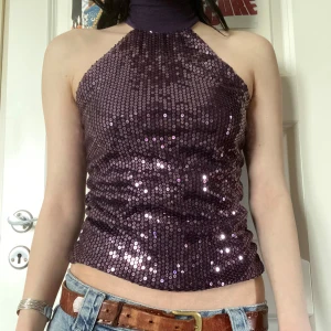 Lila paljett-topp med halterneck - Säljer en glittrig lila halterneck-topp täckt av paljetter. Toppen har hög hals och är ärmlös, perfekt för en fest eller en y2k look! Snygg att styla med jeans eller kjol. Älskar denna toppen men passar mig inte längre😭