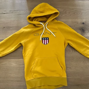Gul hoodie från GANT med logga - Säljer en gul hoodie från GANT med broderad logga och emblem på bröstet. Tröjan har huva med vita snören och en stor magficka framtill. Perfekt för dig som gillar sportig och avslappnad stil.