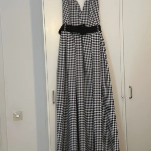 Svartvit långklänning med bälte - Snygg långklänning i svartvitt rutigt mönster med v-ringning och brett svart bälte i midjan. Klänningen är ärmlös och har en klassisk, elegant look som passar perfekt till speciella tillfällen.