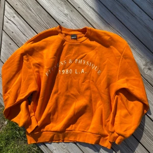 Orange sweatshirt från Gina Tricot - Säljer en orange sweatshirt från Gina Tricot med texten 'Fitness & Physique 1980 L.A.' framtill. Tröjan har rund halsringning, långa ärmar och är i mjuk bomull. Perfekt för en färgklick i garderoben!