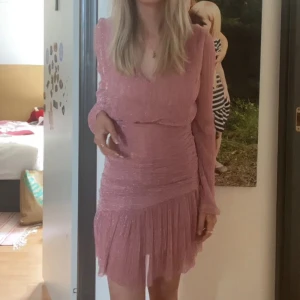 Klänning från Meet me there - Säljer en rosa kortklänning med glittrigt tyg och långa ärmar. Klänningen har en volangdetalj nertill och en lätt v-ringning. Använd några gånger men fortfarande i gott skick! 