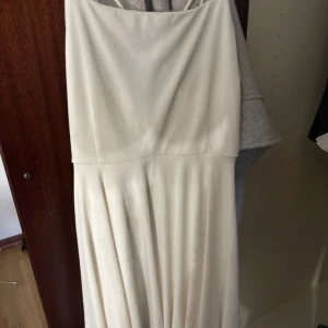 Vit långklänning med smala axelband - Säljer en elegant vit långklänning med smala axelband och mjukt fall. Klänningen har en enkel och stilren design med lätt rynkad kjol. Perfekt för sommarens speciella tillfällen eller studentfirande.