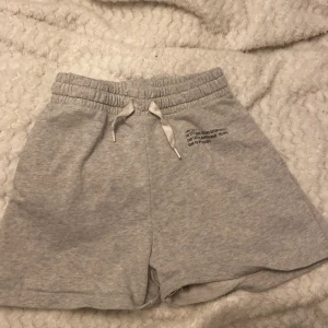 Grå mjukisshorts med tryck från H&M - Ljusgrå mjukisshorts från H&M med resår och snörning i midjan. Tryck på ena benet med text om New York. Perfekta för en avslappnad stil och sköna att ha på sig. Tillverkade i mjuk bomull.