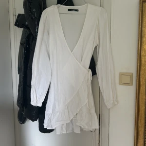 Vit omlottklänning  - Supersöt vit omlottklänning med långa ärmar och volangkant nertill. Djup v-ringning och knyt i sidan ger en snygg och feminin siluett. Perfekt för dig som gillar en fräsch och enkel stil och perfekt till student eller skolavslutning! 💌Köparen står för frakten!