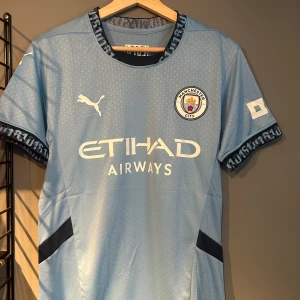Manchester City hemmatröja Puma - Manchester Citys officiella ljusblå hemmatröja från Puma med klubbmärke och Etihad Airways-tryck på bröstet. Tröjan har korta ärmar, diskret geometriskt mönster och svarta detaljer på sidorna samt textdetaljer på krage och ärmslut. Tillverkad i lätt och ventilerande material. Helt ny med tagg en replika