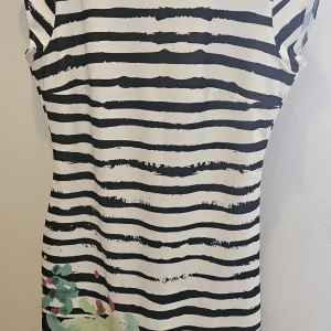 Randig klänning från Desigual XL - Snygg kortklänning från Desigual i vitt och svart med oregelbundna ränder och färgglada blommiga detaljer nertill. Klänningen har korta ärmar och en djup rundringning framtill. Perfekt för dig som gillar unika mönster och en lekfull stil.