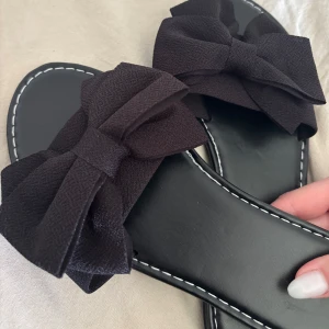 Svarta sandaler med rosetter - Stilrena svarta sandaler med breda band och stora tygrosetter framtill. Snygg kontrastsöm runt sulan och platt sula för en avslappnad vibe. Perfekta att glida runt i under varma dagar.