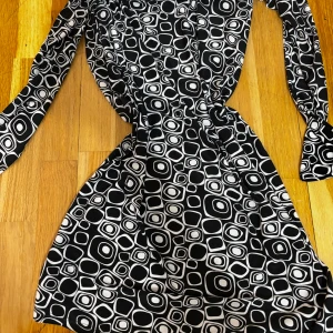 Svartvit mönstrad omlottklänning - Säljer en svartvit omlottklänning med unikt grafiskt mönster av fyrkanter och cirklar. Klänningen har lång ärm och är i mjukt, stretchigt material som sitter snyggt på kroppen. Perfekt för dig som gillar retrostil och vill sticka ut. Skirt snygg egentligen framhäver kropp öppen rygg ish