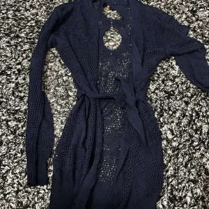 Mörkblå stickad kofta med knytband - Säljer en mörkblå, långärmad stickad kofta med knytband i midjan. Koftan har ett genomgående hålmönster och dekorativa detaljer framtill. Perfekt att slänga över en topp för en snygg lager-på-lager-look. Lite öppen i ryggen