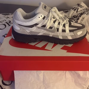Nike p-6000 - Nike p-6000 sneakers i en snygg mix av grått, vitt och svart. Skorna har mesh- och syntetpaneler, tydlig snörning och en robust, svart yttersula. Perfekta för dig som gillar sportig och modern stil.