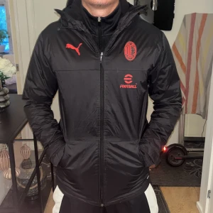 Svart AC Milan jacka från Puma - Säljer en svart jacka från Puma med AC Milan-logga och röda detaljer. Jackan har huva, dragkedja och lång ärm. Perfekt för fotbollsälskare som vill visa sitt lag. Snygg och sportig design.
