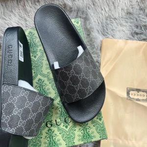 Svarta sandaler från gg - Säljer ett par svarta sandaler från Gg med klassiskt GG-mönster i grått på remmen. Tofflorna har platt sula och öppen tå, perfekta för sommaren. Kommer med originalkartong och dustbag.