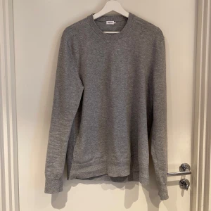 Grå stickad tröja från Filippa K - En stilren grå stickad tröja från Filippa K med rund halsringning och långa ärmar. Tröjan har en klassisk passform och är perfekt för lager-på-lager. Enkel design som funkar till många olika looks.