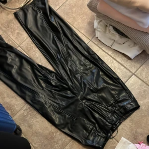 Svarta skinnbyxor från Boohoo - Snygga svarta skinnbyxor från Boohoo i storlek 36. Byxorna har raka ben, hög midja och stora fickor framtill. Tillverkade i syntetmaterial med en glansig finish som ger en cool look. Perfekta för dig som vill sticka ut med en edgy stil.