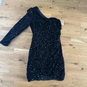 Svart paljettklänning med oneshoulder - Säljer en svart glittrig kortklänning med paljetter och oneshoulder-design. Klänningen har lång ärm på ena sidan och är figurnära med snygg drapering. Perfekt för dig som vill sticka ut på festen!