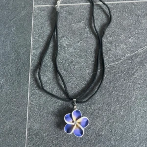 Halsband med lila blomma - Säljer ett halsband med svart band och ett hänge i form av en lila blomma med en liten pärla i mitten.  Perfekt till sommaren! Köpt utomlands men aldrig använd! 