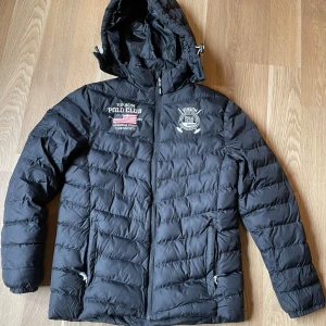 Svart pufferjacka Vinson Polo Club - Svart pufferjacka från Vinson Polo Club med loggor på bröstet och ärmen. Jackan har huva, dragkedja framtill och två fickor med dragkedja. Quiltad design och sportig look, perfekt för kalla dagar.