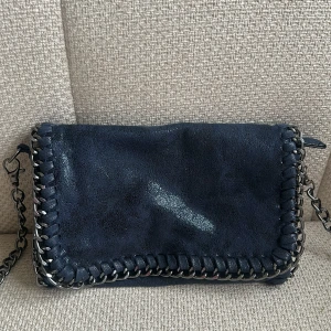 Mörkblå Tiamo axelväska med kedjedetaljer - Snygg mörkblå axelväska i mocka-look med silverfärgade kedjedetaljer runt kanterna och som axelrem. Väskan har lock med magnetknäppning och dragkedja under locket. Perfekt storlek för det viktigaste och ger en cool vibe till outfiten.